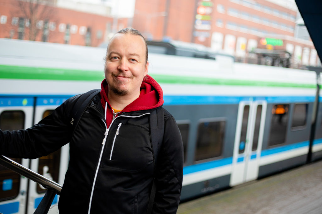 Kaajakari katsoo hymyillen kameraan, seisoo rautatieaseman portaissa. Taustalla juna.