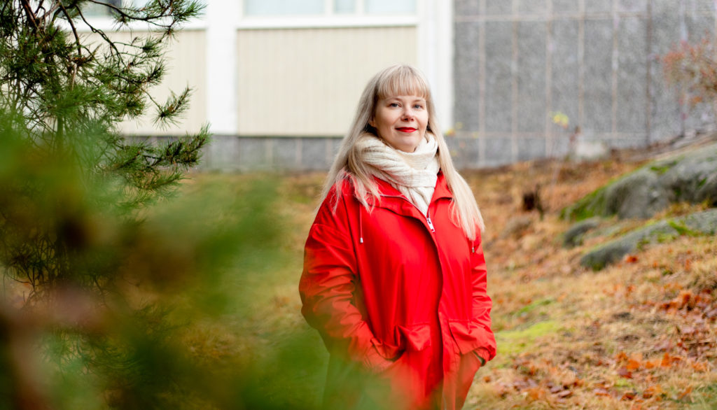 Malin katsoo kameraan hymyillen, punainen takki päällään. Etualalla havupuu, taustalla koulun seinä.